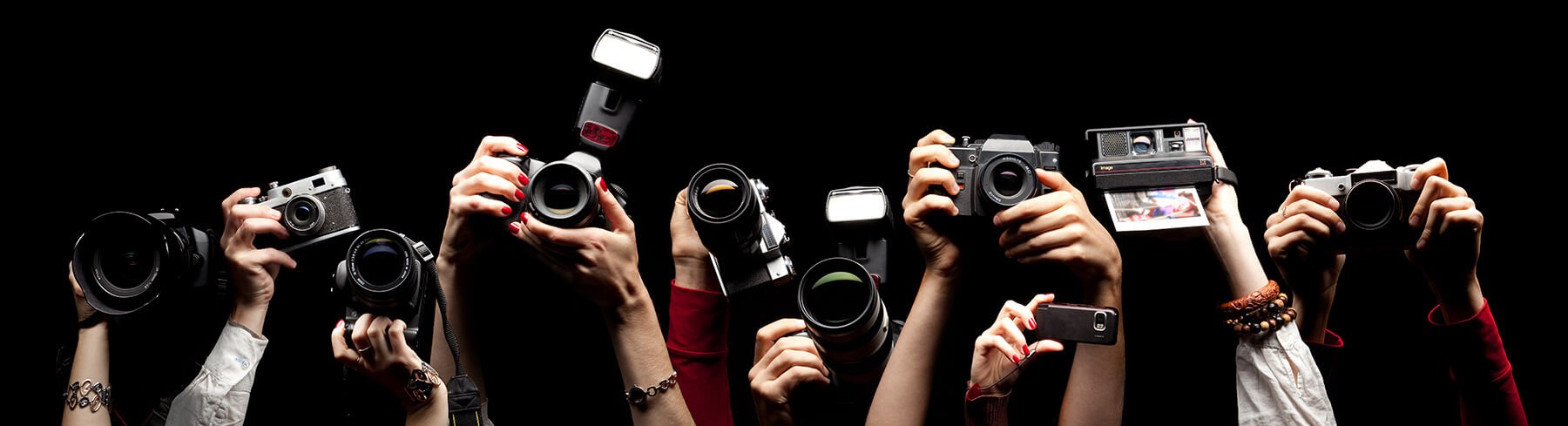 A photograph of different sorts of cameras being held up, as if at a press conference. Only the hands and forearms of the people are visible. The background is black. The image is to suggest a range of different photographers from which you have to select someone for your photoshoot.