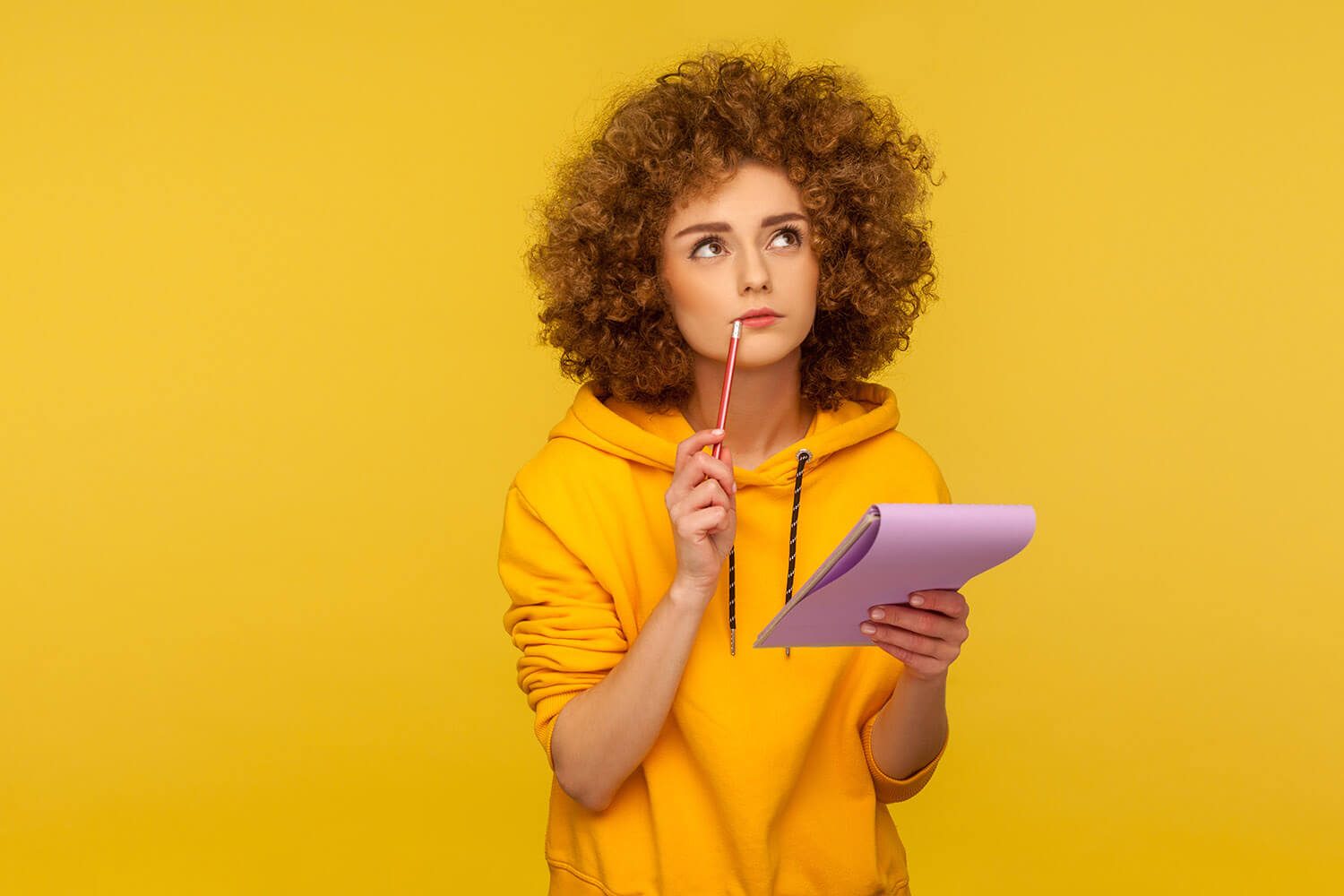 A studio photo of a young woman in a plain yellow hoodie on a plain yellow background with curly ginger hair, holding a purple note pad and a pink pencil to her lips, as if she's thinking about what to write.