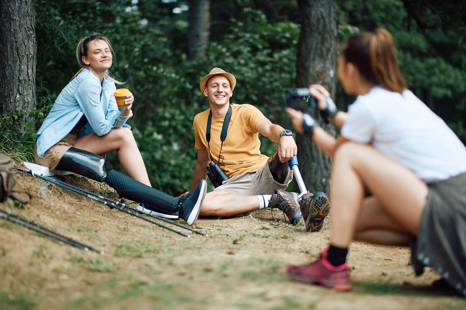 A young couple being photographed in the woods whilst on a hiking trip. There are walking poles in the image and the couple and the photographer are wearing hiking boots.