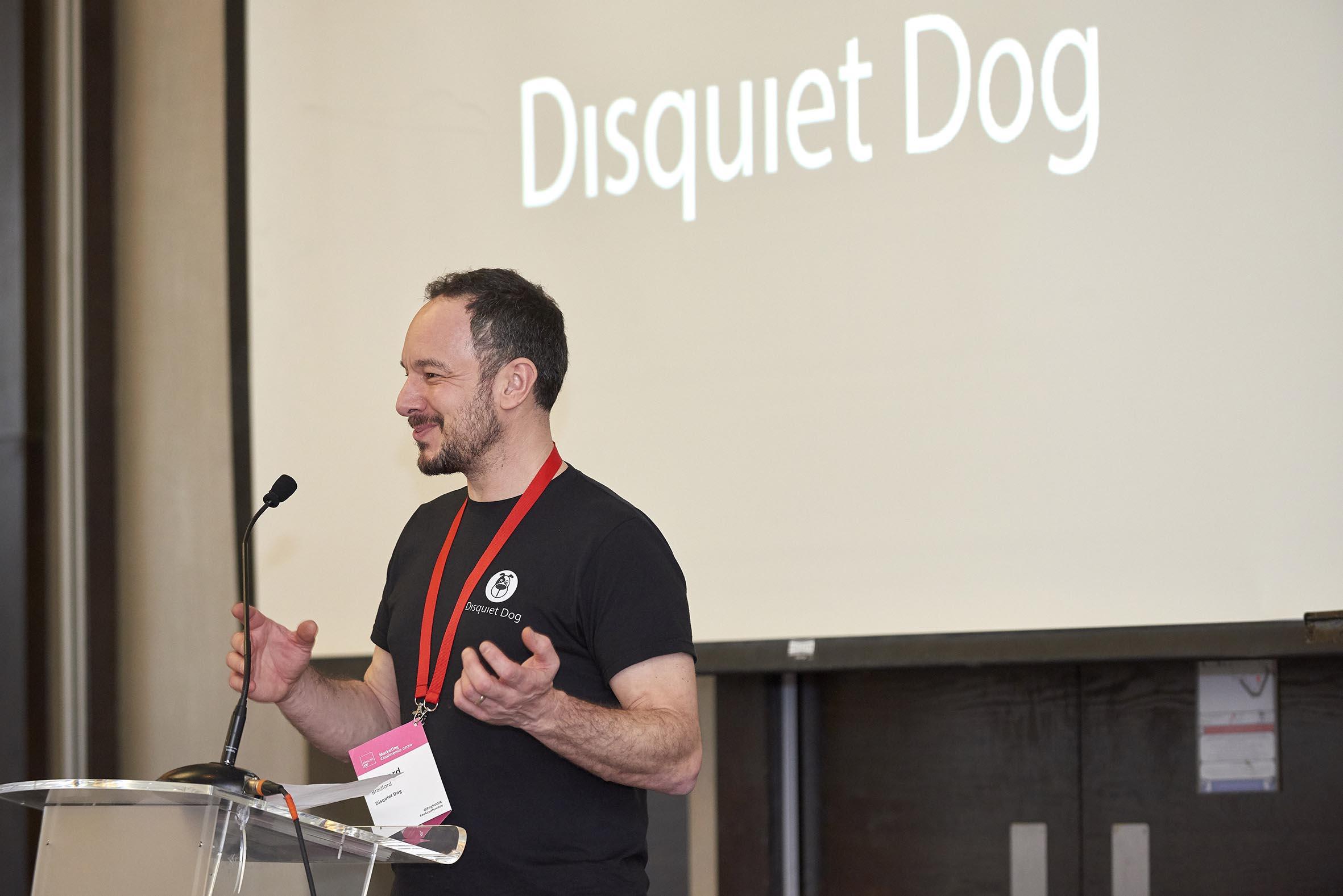 A photo of Disquiet Dog founder, Richard Bradford, speaking at a conference. He's smiling, looking at the audience which are out of the frame. Behind him are the words "Disquiet Dog" projected onto a screen. He's wearing a black T-shirt with the company's logo on it, which is the face of a dog with a black patch over one eye.