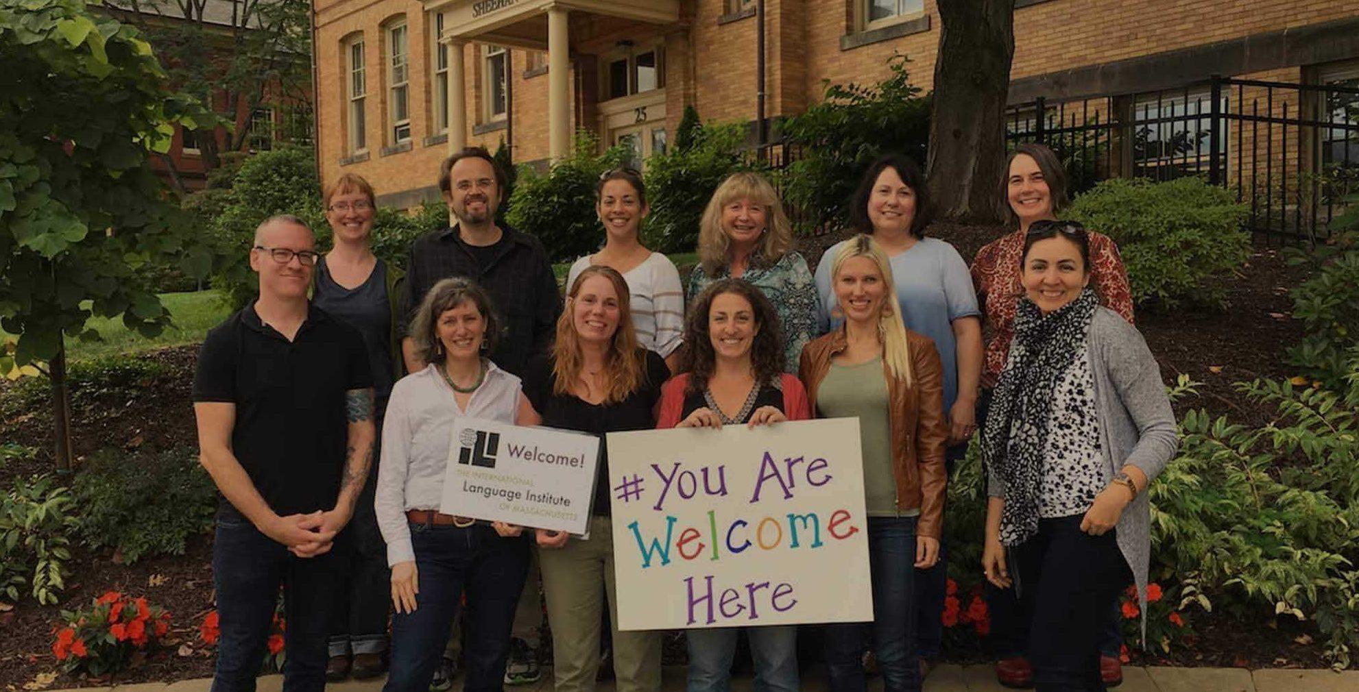 Members of the teaching team and board of ILI Massachusetts stand in front of their school, housed in an impressive brick built establishment with tall windows set in its own grounds of grass, flowerbeds and and mature trees. Some of the 12 staff are holding signs saying "You are welcome here". Everyone is smiling cheerfully.
