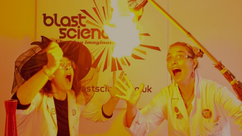 Two presenters dressed in lab coats look on excitedly at the small explosion created as a gas is set alight.