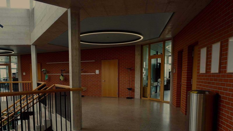 a photo of the interior upper-floor landing of a modern school building. To the left is a staircase rising up to the floor, the walls are redbrick and the colours muted.