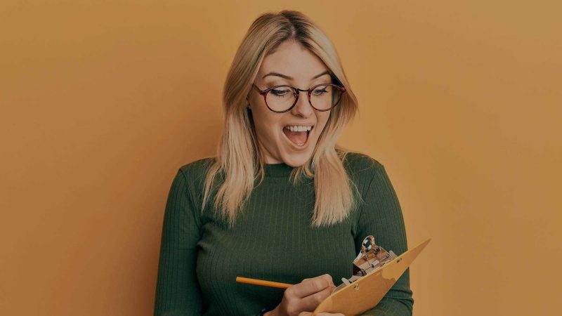 A photo of a woman in round horn-rimmed glasses with a green top and jeans, holding a yellow pencil and ocre coloured clipboard on a similar-colour background. The suggestion is that she is enthusiastically ticking something off a to do list, making progress, and she looks quite happy about it.