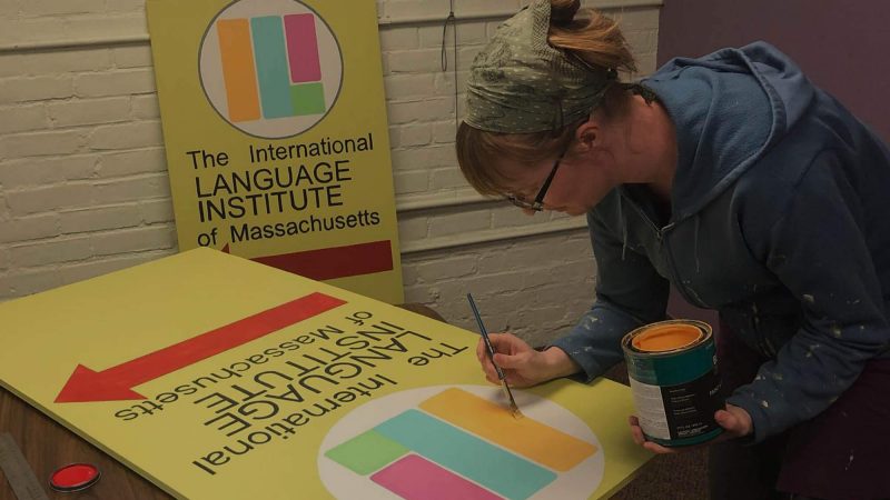 A member of the team is hand-painting a sign for the International Language Institute of Massachusetts. The sign looks like it would stand up on a sidewalk. The ILI logo is yellow, green, blue and red and the text is black on a pale yellow background.