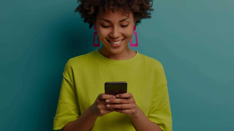 A woman with a lime green T-shirt and bright pink trapezoid shaped earrings is smiling at her mobile phone, taken here to suggest she's pleased with how the website she's viewing is performing on mobile. She's in a pale turquoise background, which makes the pink of her earrings and her T-shirt stand out.