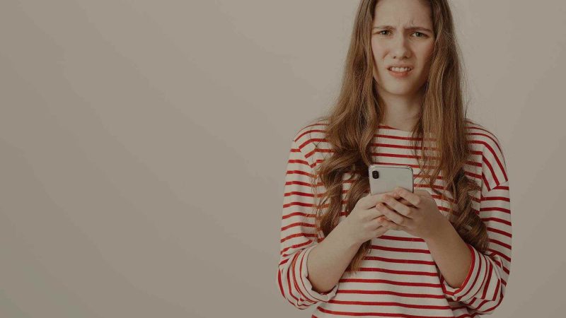 A photo of a millennial-aged girl holding a phone but looking quizzically into the camera, as if to say 