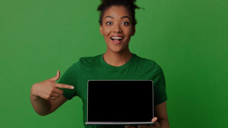 A photo of a woman smiling and pointing to a laptop enthusiastically. She's wearing a dark green T-shirt and there is a plain bright green background behind her. This photo is mildly suggestive that this article may be about websites, but it's all very figurative.