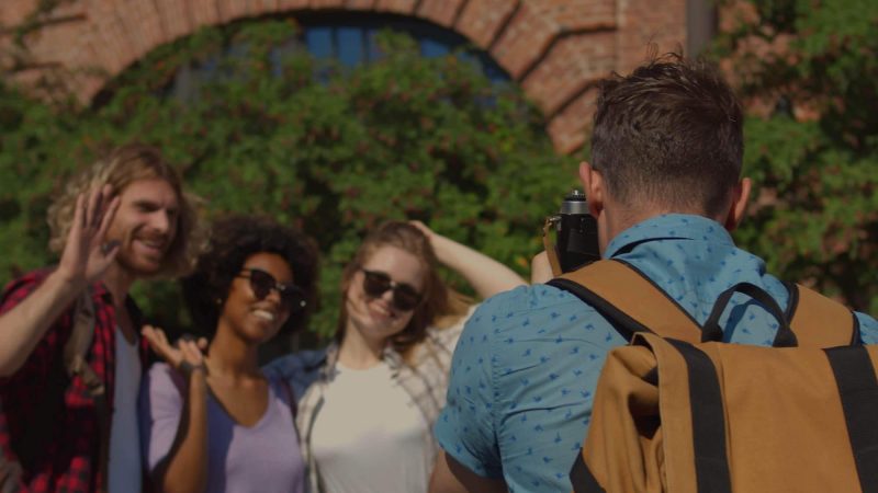 An image of a photography with his back to the camera wearing a blue shirt and a beige and black photographer's rucksack taking a picture of three people outside on a sunny day. The photo is taken in a city with a brick archway and trees slightly blurred in the background.