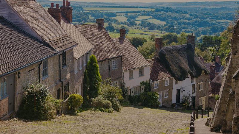 A colour photo of the view from Gold Hill in Shaftesbury, England which is often used in tourism marketing. This popular tourist spot evokes emotions of old England with a mixture of traditional terraced cottages include one thatched cottage down one side of a very steep hill. In the background you can see the English countryside disappearing to the horizon.