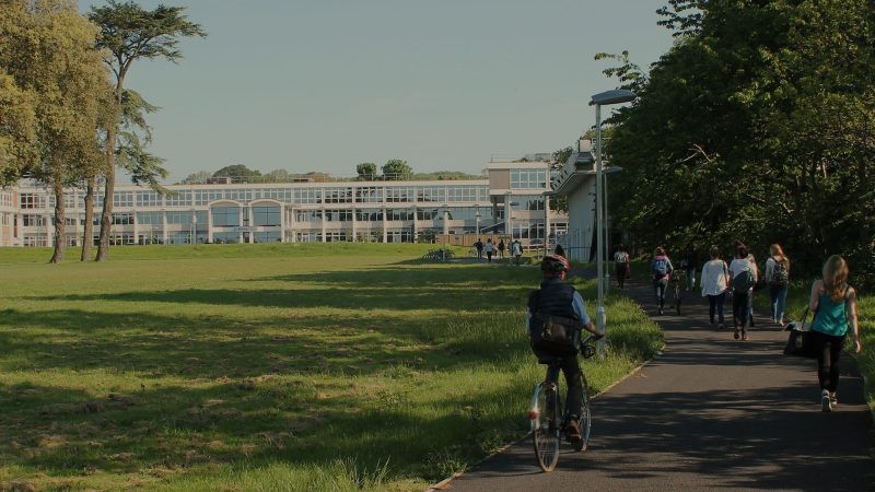 A photograph of Worthing college in the distance with fresh green lawns and tall trees in the foreground. Students can be seen walking up a path to the college.