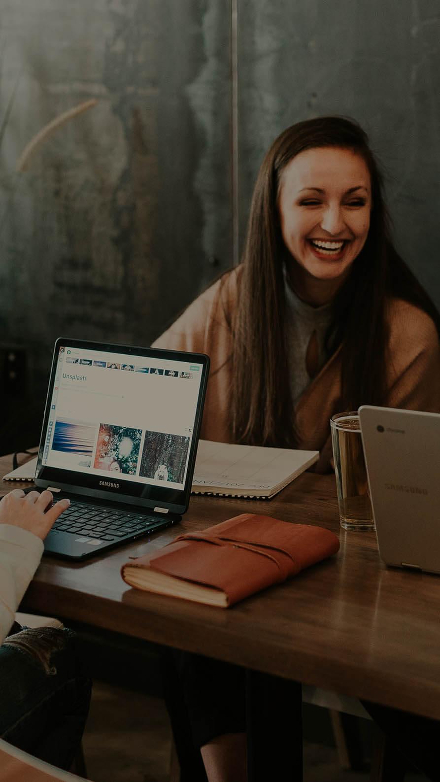 A photo of three people who might be attending the digital marketing bootcamp programme. They are sitting around a wooden table. Two of them have laptops open. They are laughing and seem to be having a good time.