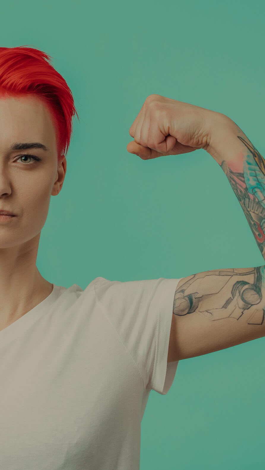 An image of a strong female body builder with short, bright red hair looking straight into the camera lens with a deterimined look in her eyes. She's wearing a white T-shirt which exposes a colourful sleeve tattoo. She is tensing her bicep to show her muscles.