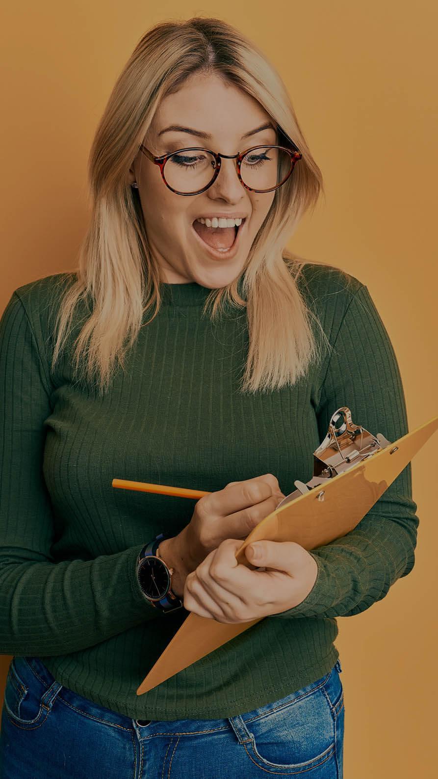 A photo of a woman in round horn-rimmed glasses with a green top and jeans, holding a yellow pencil and ocre coloured clipboard on a similar-colour background. The suggestion is that she is enthusiastically ticking something off a to do list, making progress, and she looks quite happy about it.