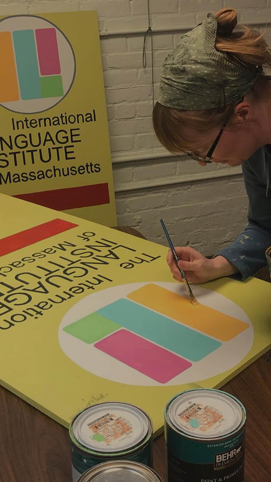A member of the team is hand-painting a sign for the International Language Institute of Massachusetts. The sign looks like it would stand up on a sidewalk. The ILI logo is yellow, green, blue and red and the text is black on a pale yellow background.