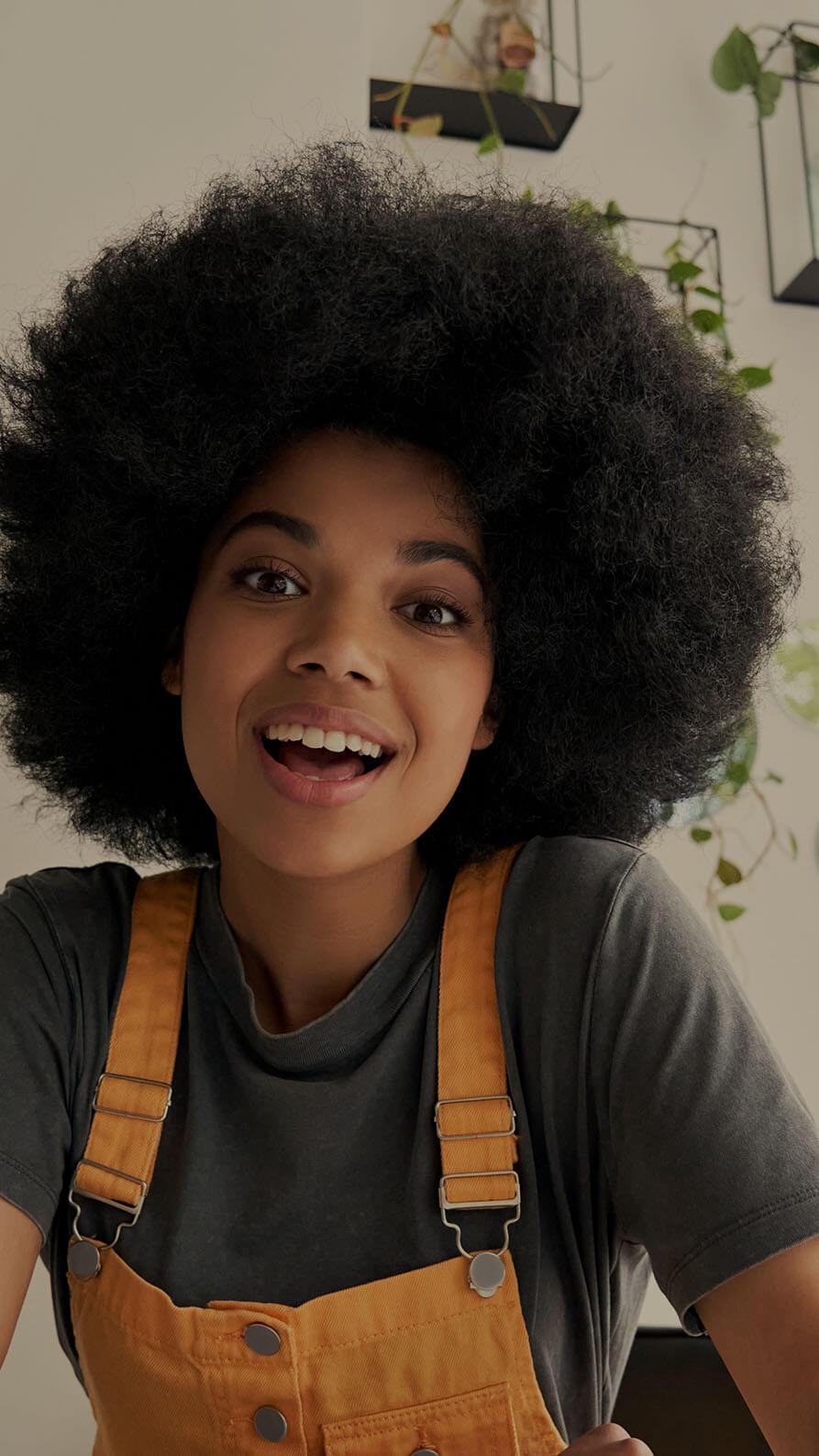 A young woman with a big smile, speaking energetically presumably into a camera. We think she's a social media influencer. She's wearing a grey T-shirt, and orange dungarees. In the background is a white wall with a selection of plants growing down from several small wall-mounted wire and glass terrariums.