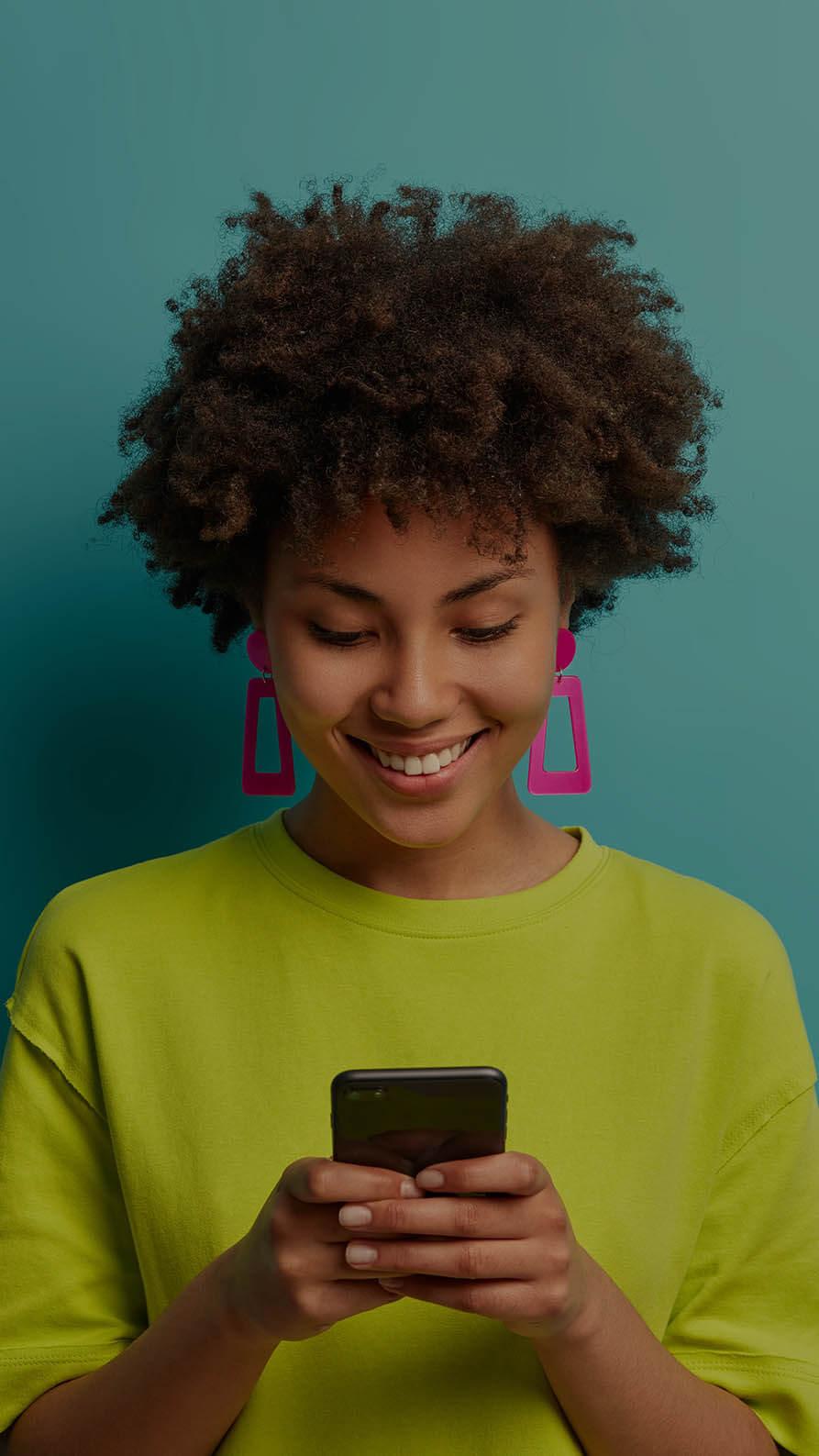 A woman with a lime green T-shirt and bright pink trapezoid shaped earrings is smiling at her mobile phone, taken here to suggest she's pleased with how the website she's viewing is performing on mobile. She's in a pale turquoise background, which makes the pink of her earrings and her T-shirt stand out.