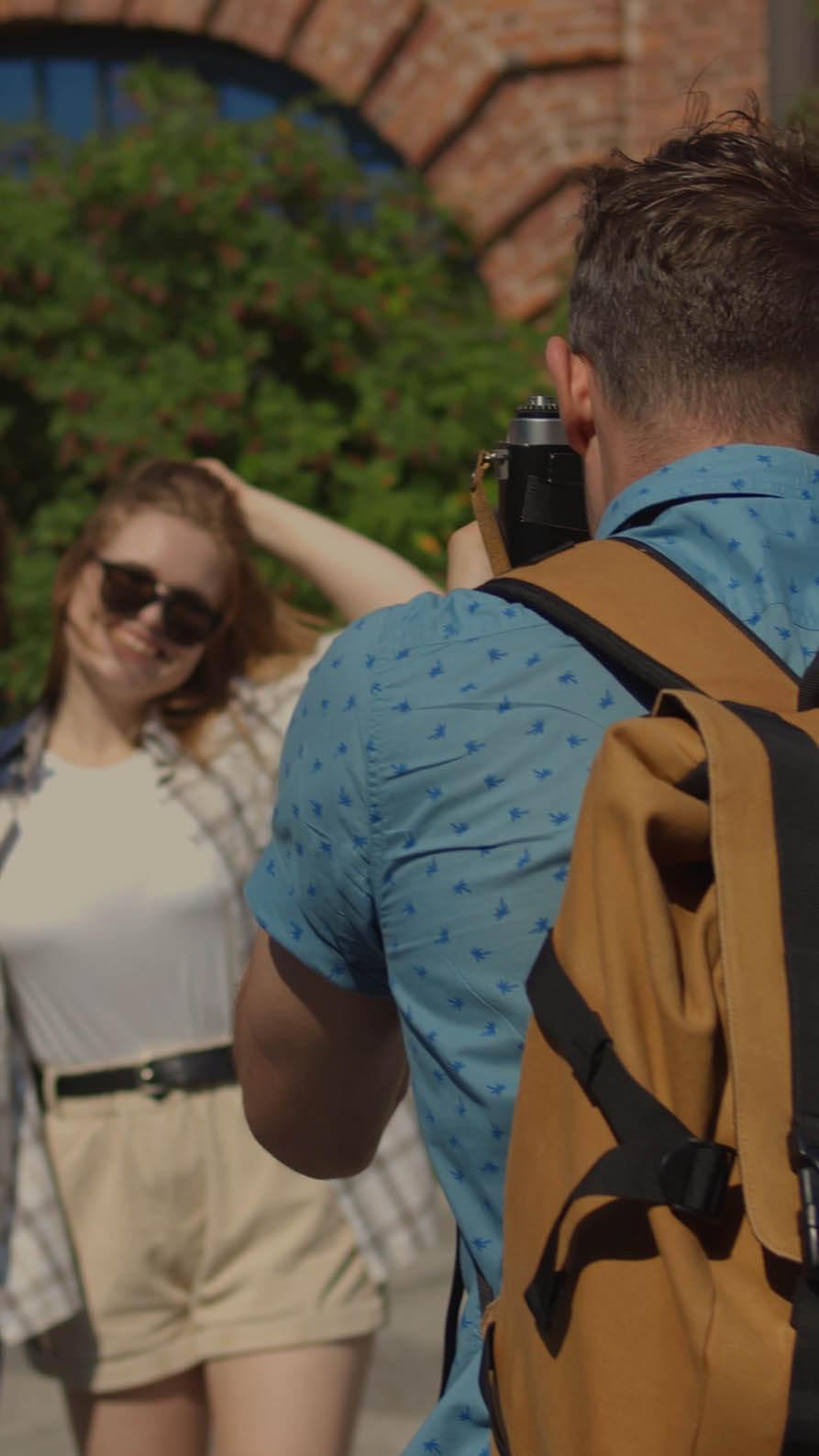 An image of a photography with his back to the camera wearing a blue shirt and a beige and black photographer's rucksack taking a picture of three people outside on a sunny day. The photo is taken in a city with a brick archway and trees slightly blurred in the background.