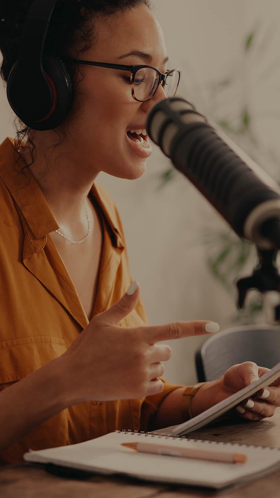 A photo of a young woman wearing an burnt orange coloured blouse speaking into a microphone whilst glancing down at her notes. She's wearing headphones and we think she might be recording a podcast. She's sitting at a wooden table, and there's a plant, blurred, in the background.