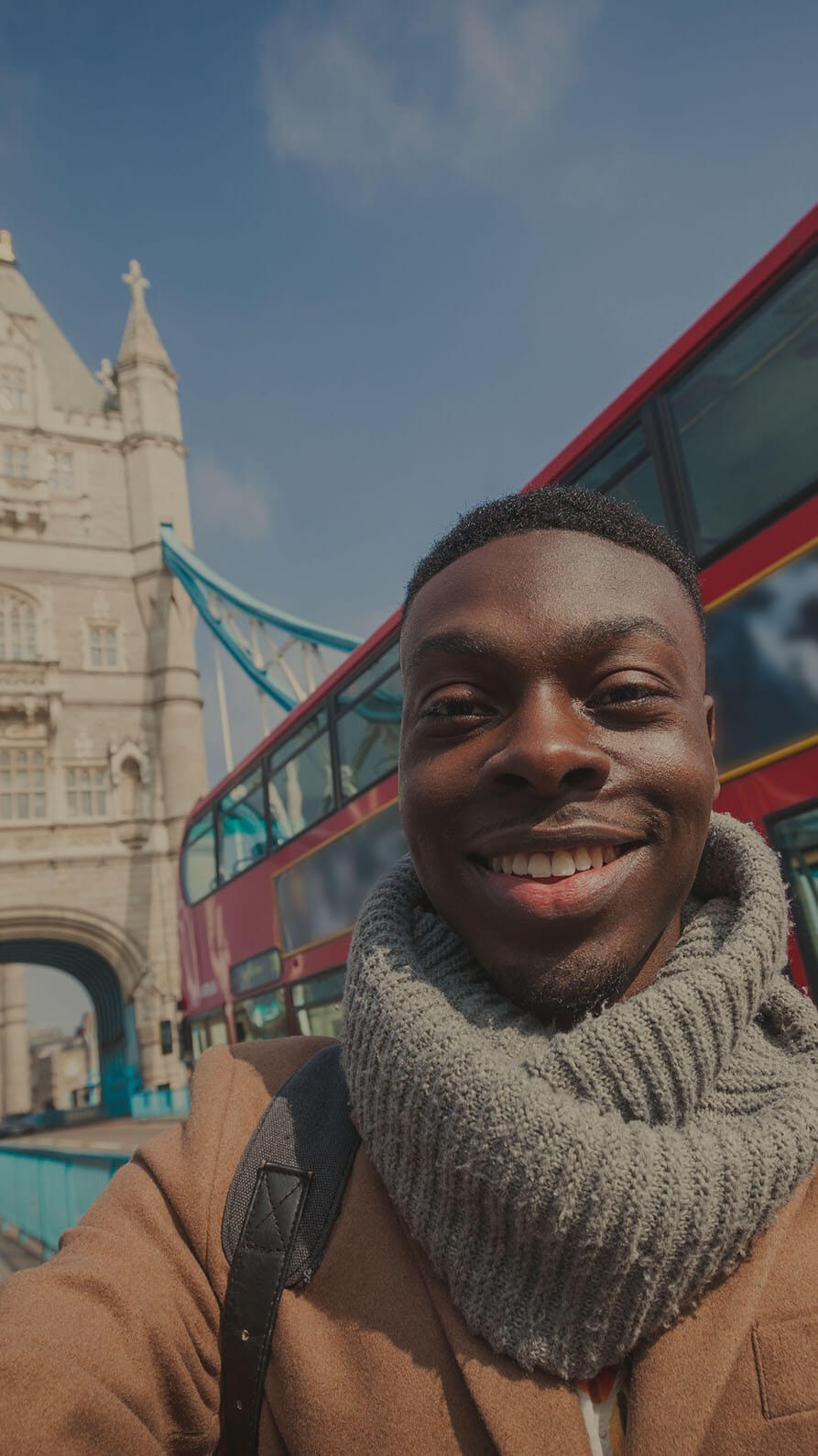 A smiley young man wearing a grey scarf and smart brown coat is posing for a selfie in front of one of the towers of Tower Bridge in London, whilst a red london double decker bus passes behind him. It looks like he's a tourist or a social influencer.