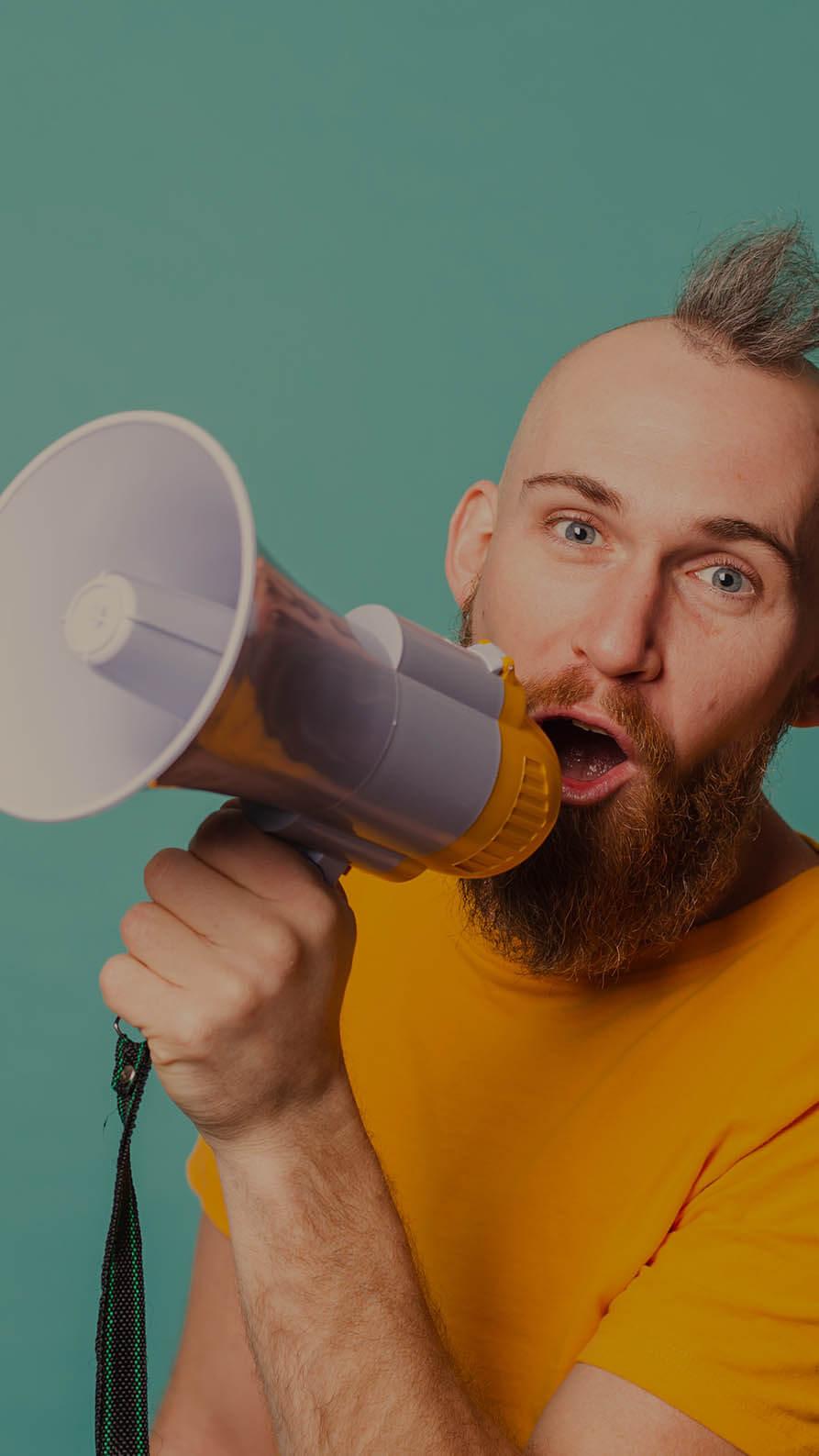 A man with a beard and mohican hair style wearing a yellow T-shirt is looking directly into the camera whilst holding a yellow and white megaphone to his mouth. He's pictured on a plain turquoise background. The image  is being used to suggest a casual tone of voice in corporate communications.