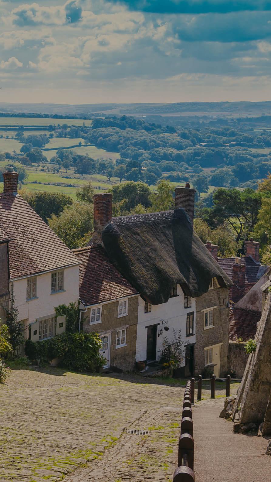A colour photo of the view from Gold Hill in Shaftesbury, England which is often used in tourism marketing. This popular tourist spot evokes emotions of old England with a mixture of traditional terraced cottages include one thatched cottage down one side of a very steep hill. In the background you can see the English countryside disappearing to the horizon.